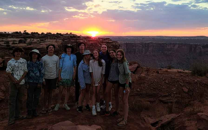 A group of students smile for a group photo while the sun sets behind them.